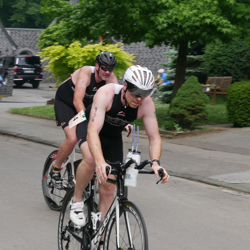 Zwei Triathleten fahren auf Rennrädern eine Straße entlang, während ein dritter Radfahrer im Hintergrund sichtbar ist.