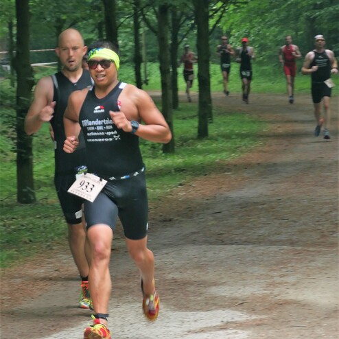 Zwei Männer beim Laufen in einem Wald, einer trägt Sonnenbrille und Stirnband, andere Läufer im Hintergrund.