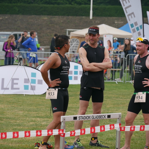 Drei Triathleten in Wettkampfkleidung stehen im Gespräch vor einer Sportveranstaltung mit Kulisse und Zuschauern.