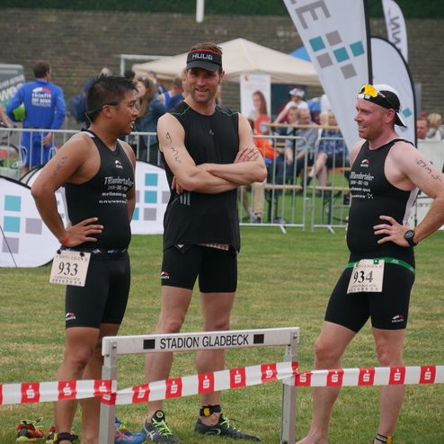 Drei Sportler in Wettkampfkleidung stehen am Rande eines Triathlons und unterhalten sich vor einer Ziellinie.