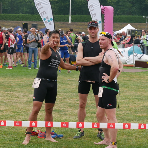 Drei Triathleten in Sportkleidung stehen auf einer Wiese, während im Hintergrund andere Teilnehmer warten.
