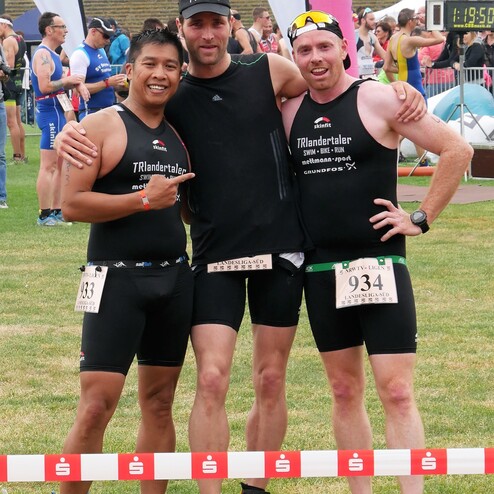 Drei Triathleten posieren lächelnd nach dem Wettkampf auf einer Rasenfläche, umgeben von Veranstaltungsflaggen.