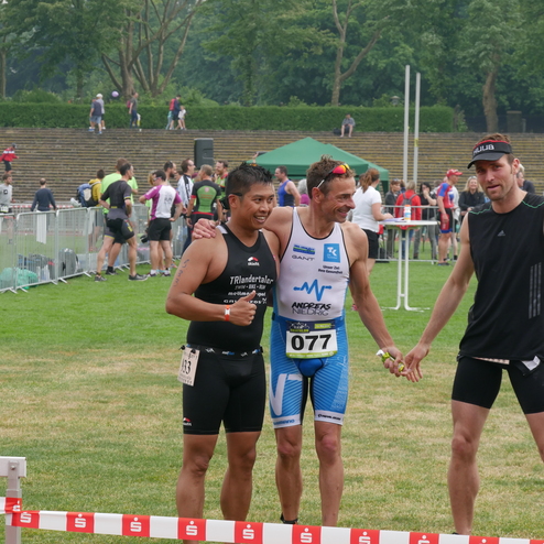 Drei Triathleten posieren nach dem Wettkampf auf einer Wiese vor Zuschauern und Absperrungen.