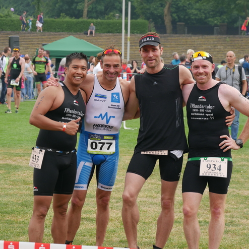 Vier Triathleten posieren an einem Wettkampftag auf einer Wiese, umgeben von Zuschauern und Event-Equipment.
