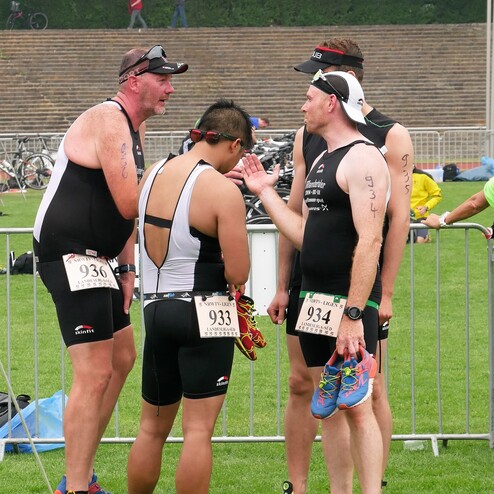 Vier Athleten in Wettkampfanzügen unterhalten sich auf einem Sportplatz, während Fahrräder im Hintergrund geparkt sind.