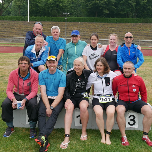 Gruppe von zwölf Sportlern in Wettkampfkleidung sitzt auf einem Podest im Freien, umgeben von Rasen und einem Laufbahn.