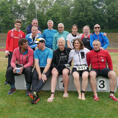 Gruppe von 15 Personen in Sportkleidung, sitzend und stehend auf einer Siegerehrungsplattform im Freien.