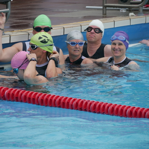 Gruppenschwimmen von sechs Frauen mit Schwimmkappen und Brillen in einem Pool, lächelnd und nah beieinander.