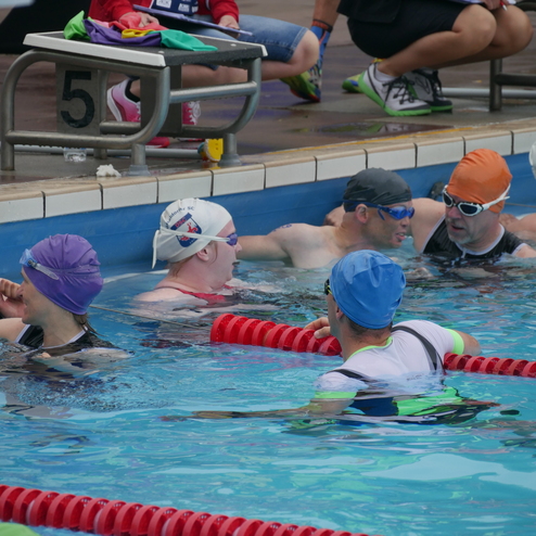 Mehrere Schwimmer in Badekappen und Schwimmbrillen unterhalten sich im Wasser eines Schwimmbeckens, umgeben von Startblöcken.
