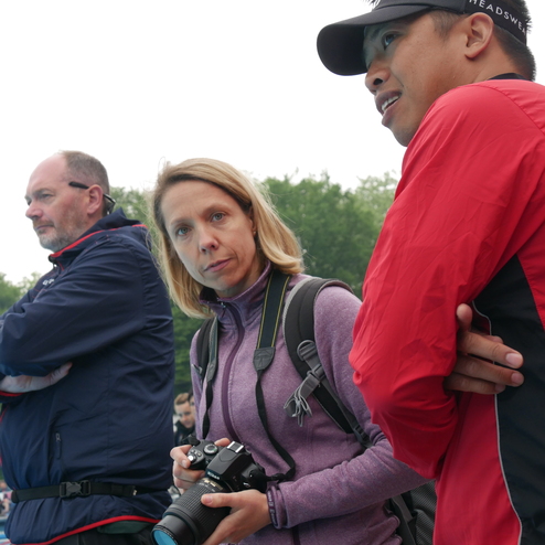 Drei Personen am Rande eines Schwimmbeckens, eine Frau hält eine Kamera, alle blicken ernst in die Ferne.