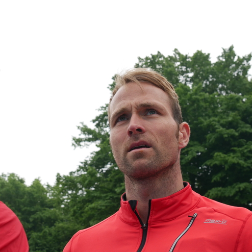 Ein Mann in roter Sportjacke schaut nach oben, Hintergrund mit Bäumen und Sportplatzbeleuchtung.