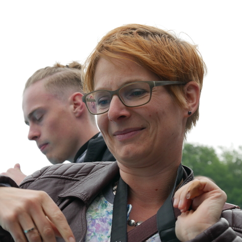 Frau mit kurzem, blondem Haar und Brille, lächelt während sie auf eine Kamera vorbereitet. Im Hintergrund sind weitere Personen.