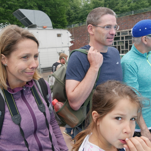 Gruppe von drei Personen, darunter ein Mädchen, stehen entspannt im Freien vor grüner Kulisse.