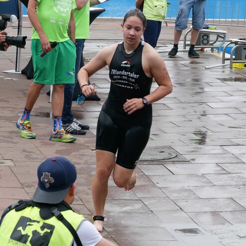 Eine Triathletin in Neoprenanzug läuft auf einem Wettkampfareal, während Zuschauer und Offizielle beobachten.