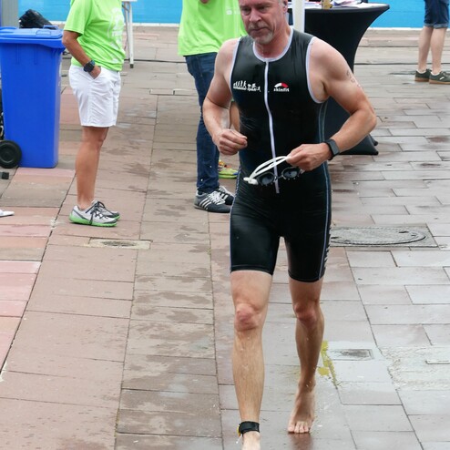 Ein Mann in einem schwarzen Triathlonanzug läuft barfuß auf einem feuchten Boden, während Zuschauer im Hintergrund stehen.