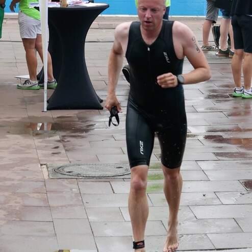 Ein Teilnehmer läuft barfuß und in schwarzem Neoprenanzug auf einem nassen Weg neben einem Schwimmbecken.