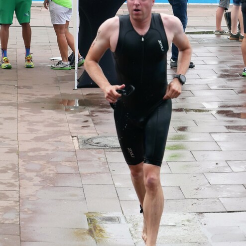 Mann in einem schwarzen Triathlonanzug läuft barfuß auf nassem Boden, umgeben von Veranstaltungsteilnehmern.