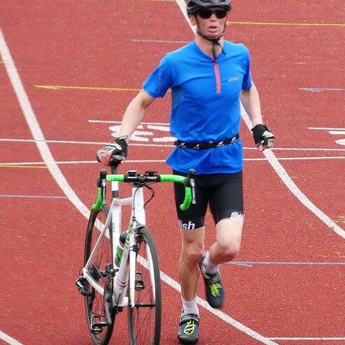 Sportler in blauer Oberbekleidung joggt mit Rennrad auf einer roten Leichtathletikbahn.