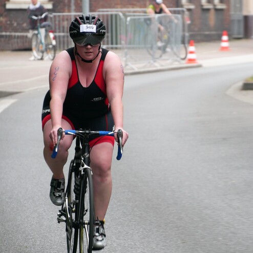 Sportlerin in schwarzer und roter Bekleidung fährt auf Straßenrad durch eine Stadt, trägt Helm mit Nummer 940.