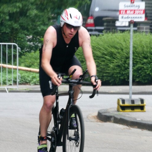 Ein Triathlet in schwarzem Anzug fährt konzentriert auf einem Rennrad entlang einer grünen Allee.