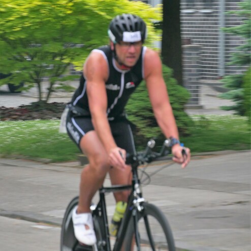 Radfahrer in sportlicher Bekleidung fährt auf einem schwarzen Fahrrad. Im Hintergrund Bäume und Wohngebäude.