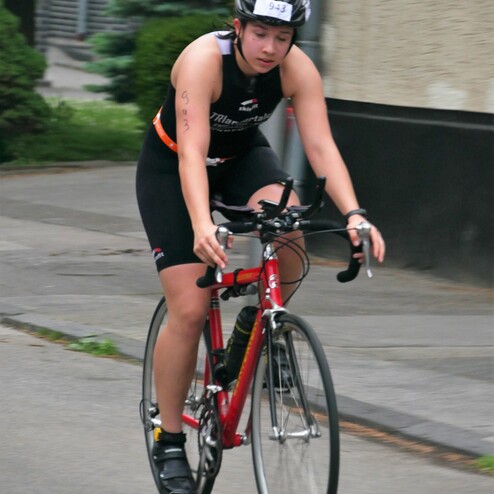 Athletin in schwarzem Sportanzug fährt auf rotem Rennrad eine Straße entlang, mit Nummernschild auf dem Helm.