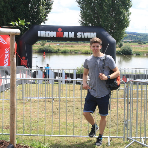 Junger Mann steht vor einem Ironman-Schwimm-Banner, im Hintergrund ist ein Gewässer und Wettkampfbereich sichtbar.
