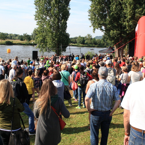Große Menschenmenge am Ufer eines Flusses, Zuschauer beobachten ein Event mit roten und gelben Markierungen im Hintergrund.
