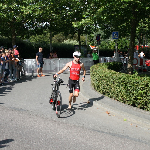 Triathlet in Rot trägt sein Fahrrad während des Rennens, umgeben von zuschauenden Menschen und Grüntönen.