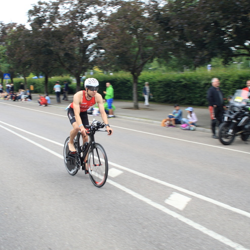 Triathlet in Sportkleidung fährt auf einem Rennrad eine Straße entlang, Zuschauer sitzen am Rand.
