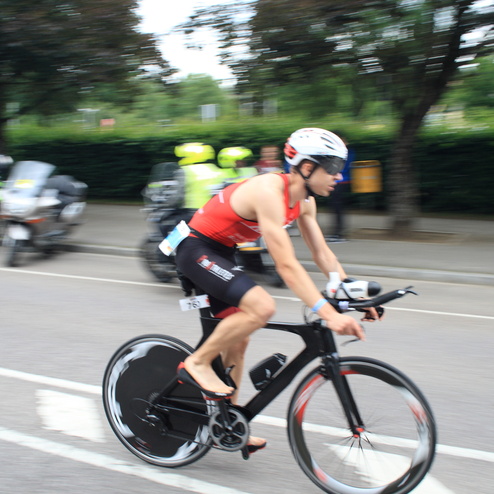 Athlet in rotem Trikot fährt mit einem Rennrad auf einer Straße, während Zuschauer und Motorräder im Hintergrund zu sehen sind.