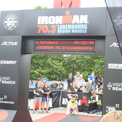 Zielbogen des IRONMAN 70.3 Luxembourg mit Zeit und Teilnehmername, umgeben von Zuschauern und Kameraleuten.