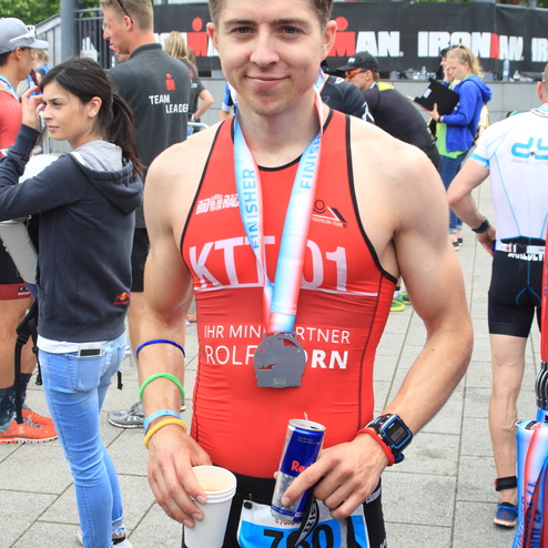 Junger Mann in Wettkampfanzug mit Medaille, Becher und Energydrink nach Triathlon-Wettbewerb, lächelt in die Kamera.