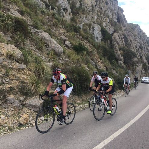 Vier Radfahrer auf einer kurvigen Straße entlang steiler Felsen, mit Fahrzeug im Hintergrund.