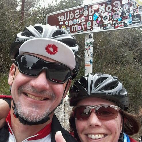 Selbstporträt von zwei Radfahrern mit Helmen vor einem mit Aufklebern bedeckten Schild auf einer Waldstraße.