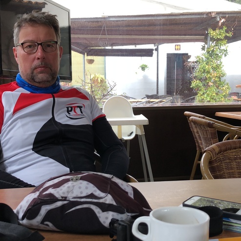 Mann in Radbekleidung sitzt am Tisch in einem Café, umgeben von Fahrradzubehör und einer Tasse Kaffee.