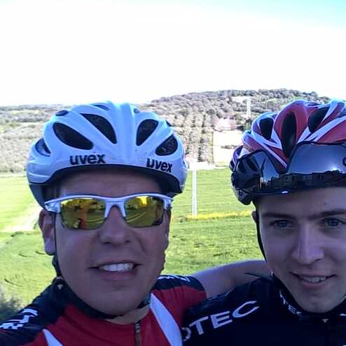 Selbstporträt zweier Radfahrer mit Helmen, vor einer grünen Landschaft mit Bäumen und Wiesen.