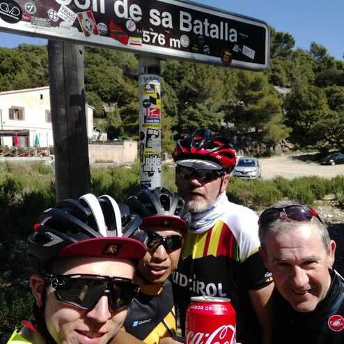 Gruppe von Radfahrern posiert am Schild „Coll de sa Batalla“ mit der Angabe 576 m, einer Dose Coca-Cola in der Hand.