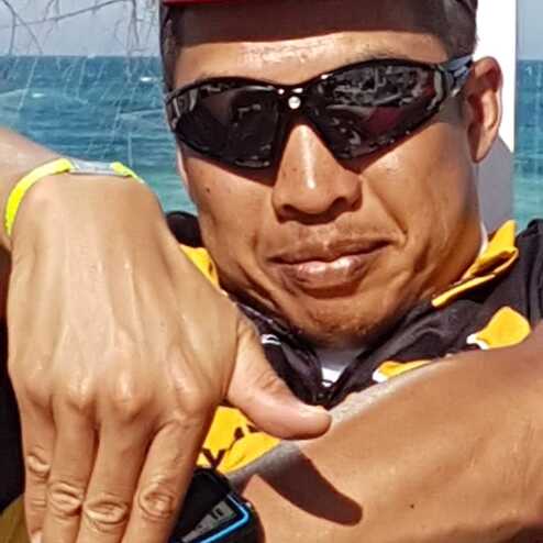 Mann in Sonnenbrille und Kapuzenjacke zeigt Handzeichen am Strand mit Wasser im Hintergrund.