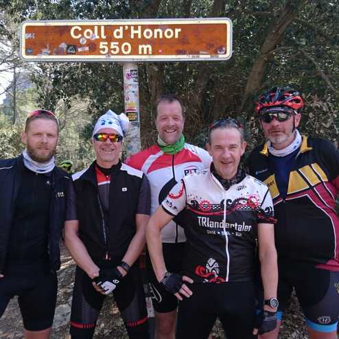 Fünf Radfahrer posieren an einem Schild für den Coll d’Honor, 550 m, umgeben von Bäumen und einer Straße.
