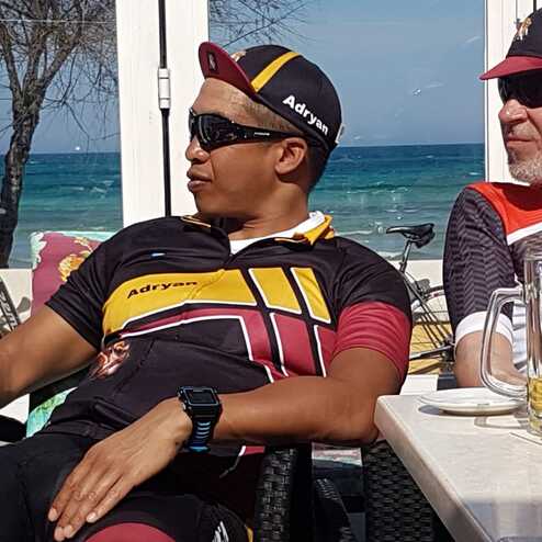 Zwei Männer in Radbekleidung sitzen an einem Tisch, mit Blick auf das Meer und Fahrrädern im Hintergrund.