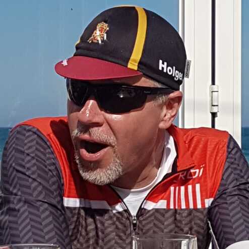 Mann mit Sonnenbrille und schwarz-roter Mütze, sitzt an einem Tisch mit Biergläsern, lächelt und spricht leidenschaftlich.