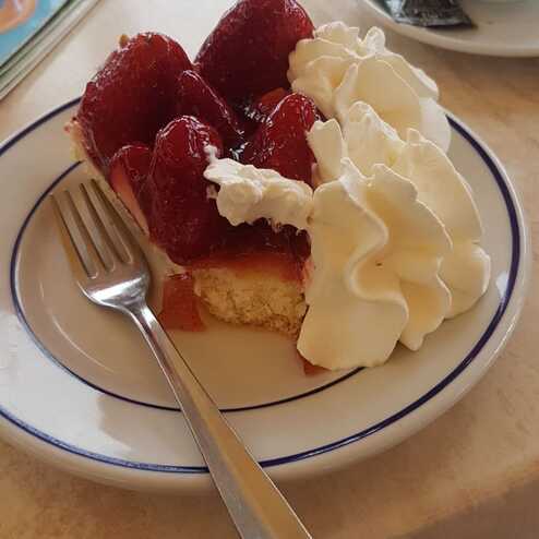 Stück Erdbeerkuchen mit Sahnehaube auf einem Teller, neben einer Gabel und einer Tasse Kaffee.