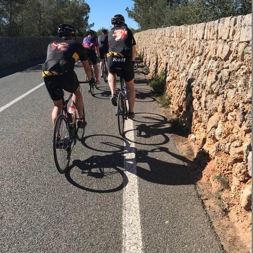 Gruppe von Radfahrern auf einer Landstraße, neben einer Steinmauer, unter blauem Himmel mit Bäumen im Hintergrund.