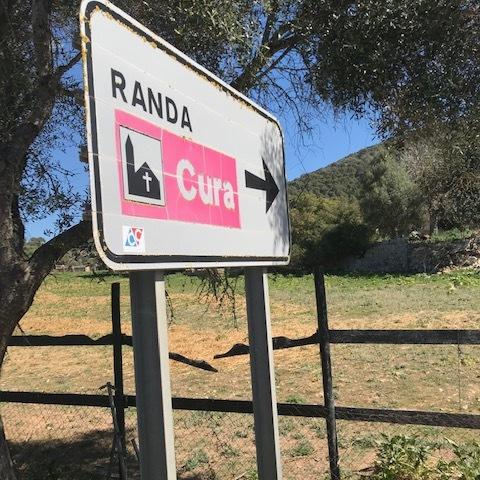 Wegweiser nach Randa und Cura mit Pfeil nach rechts, umgeben von grüner Landschaft und Bäumen.