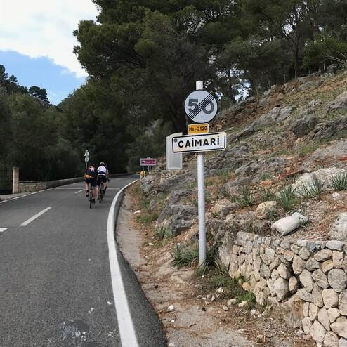 Zwei Radfahrer fahren die Straße entlang, die zum Ortsschild "Caimari" und einer Geschwindigkeit von 50 km/h führt.