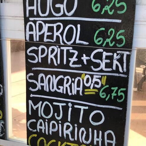 Getränkekarte mit Preisen: Hugo, Aperol, Spritz+Sekt, Sangria, Mojito und Caipirinha auf einer schwarzen Tafel.