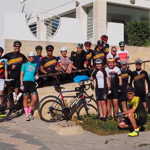 Gruppe von Radfahrern in Sportkleidung posiert lächelnd vor einem modernen Gebäude, Fahrräder neben ihnen abgestellt.