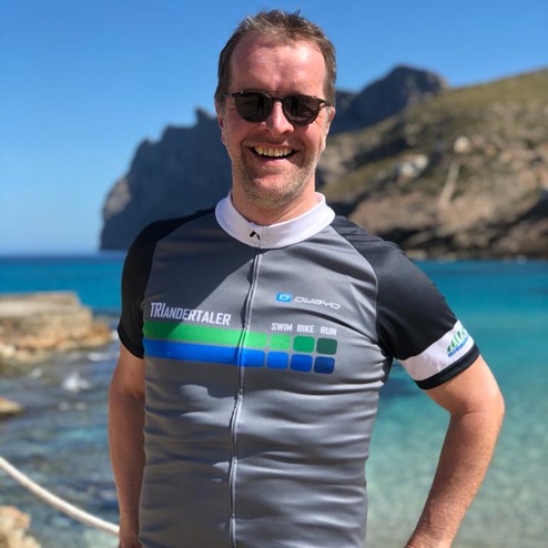 Mann mit Sonnenbrille und Sporttrikot vor einer Küstenlandschaft mit blauem Wasser und Bergen im Hintergrund.
