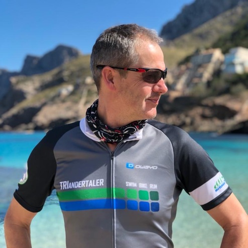 Mann in Triathlon-Outfit mit Sonnenbrille und Halstuch steht am Strand vor klar blauem Wasser und Bergen im Hintergrund.